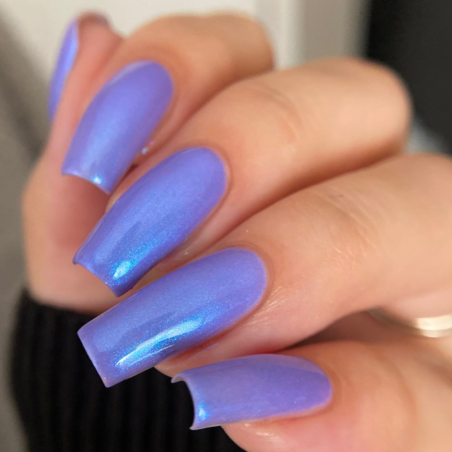 A close-up of a hand with long, rounded nails painted in a shimmering lavender-purple holographic nail polish, wearing a black ribbed sweater.
