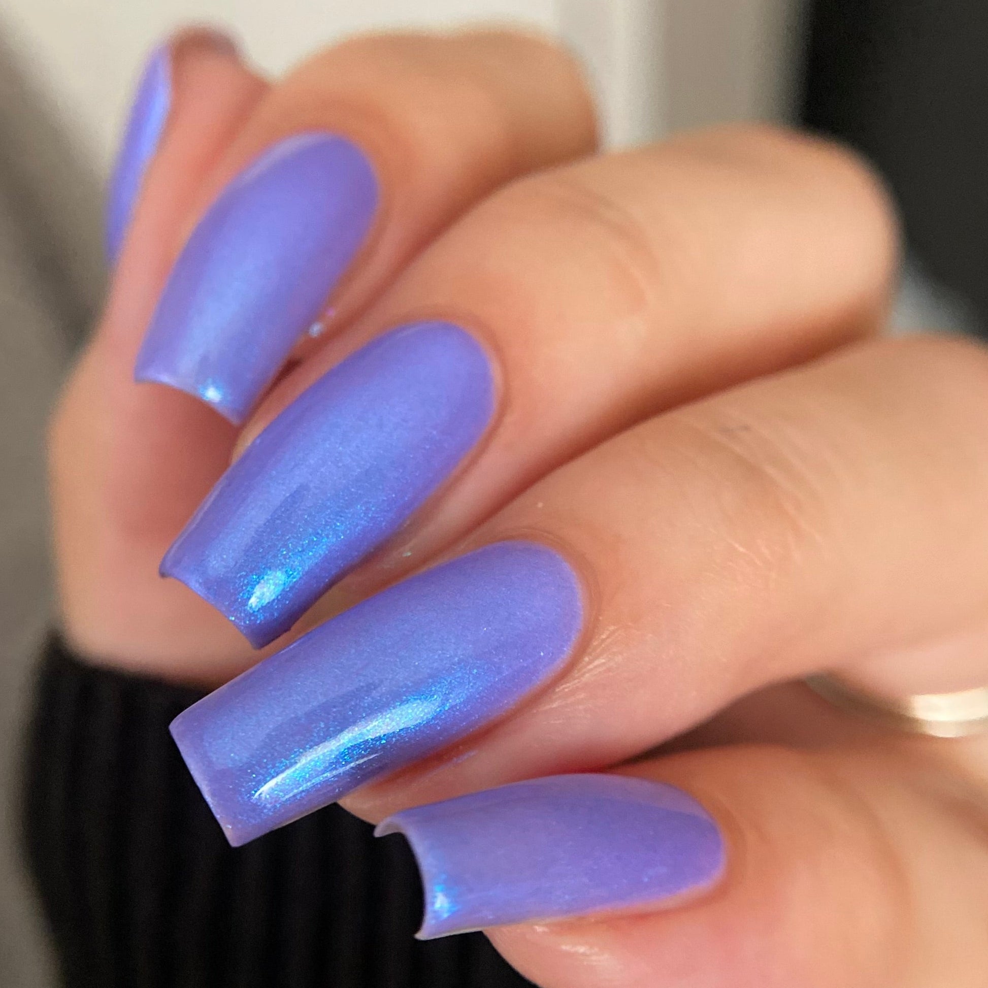 A close-up of a hand with long, rounded nails painted in a shimmering lavender-purple holographic nail polish, wearing a black ribbed sweater.