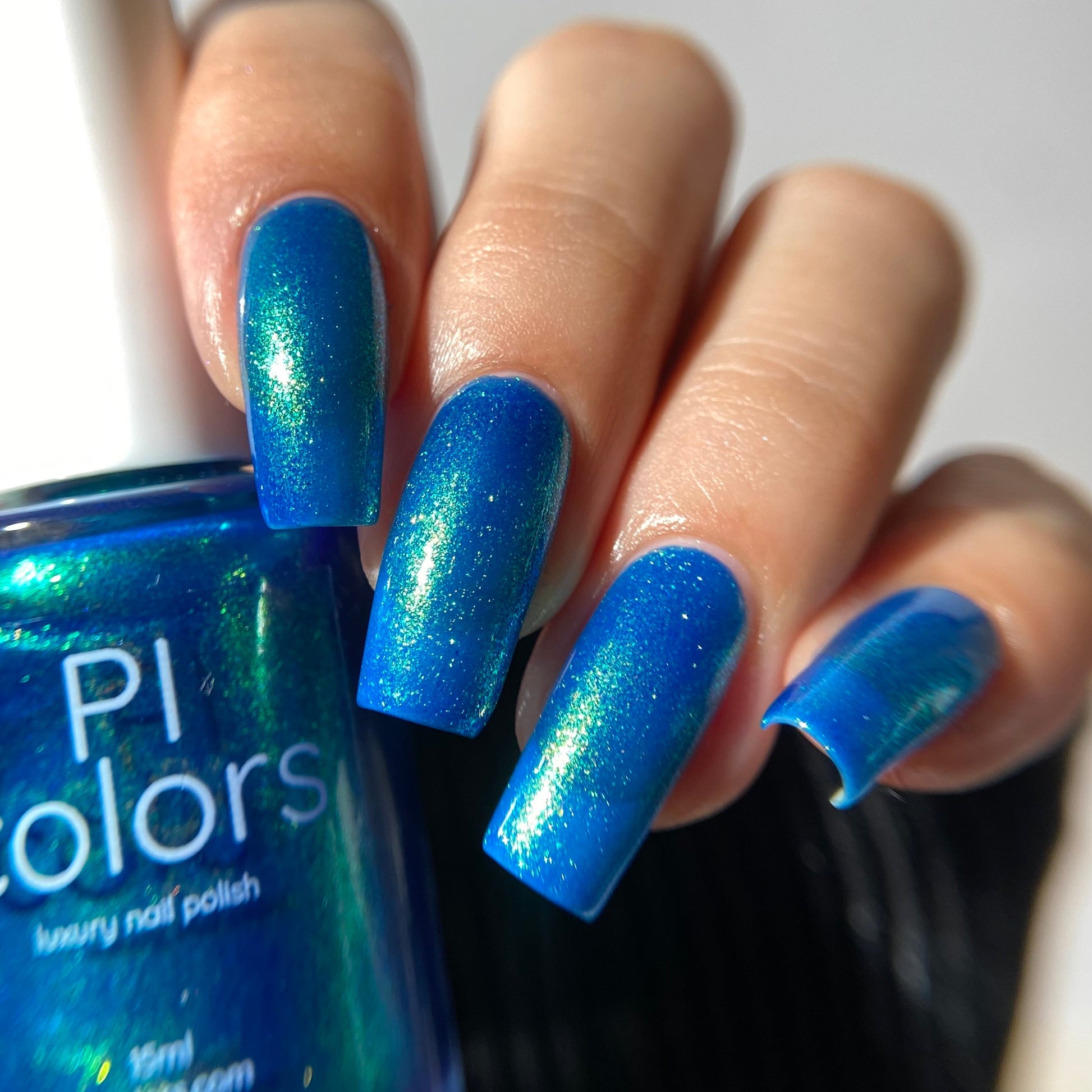 A close-up of a hand with long, glossy blue nails painted with a shimmering blue nail polish, holding a bottle of PI colors nail polish.