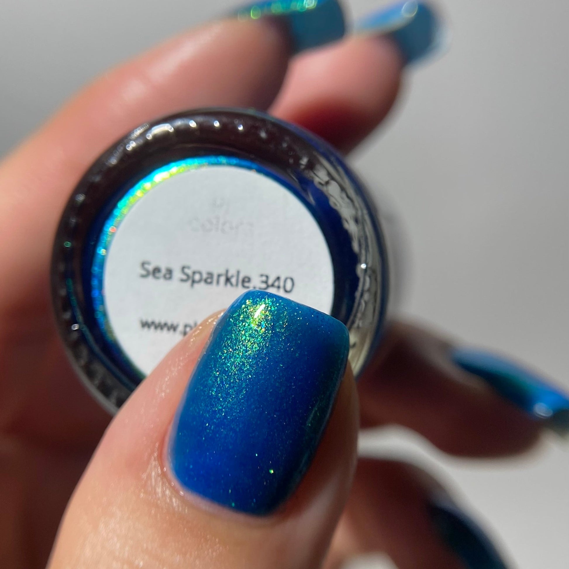 A close-up of a hand holding a small round bottle of nail polish. The visible product color is a shimmering royal blue with iridescent sparkle, and the label reads 'Sea Sparkle 340'.