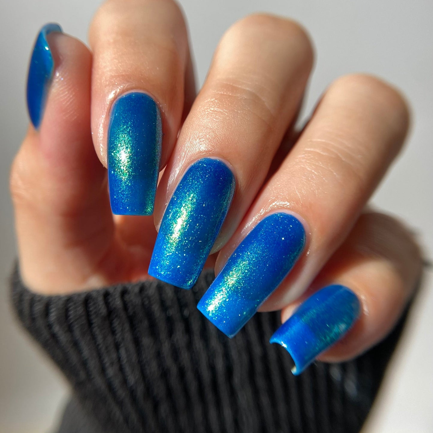 A close-up of a hand with long, almond-shaped nails painted in a vibrant blue glitter nail polish, wearing a black ribbed knit sleeve against a light gray background.
