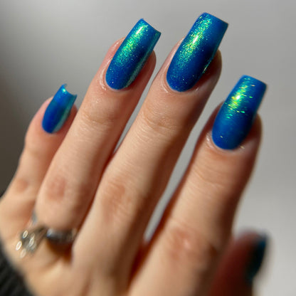 A close-up of a hand with long, square-shaped nails painted in a shimmering metallic blue nail polish with green iridescent flecks, wearing a silver ring against a soft gray background.