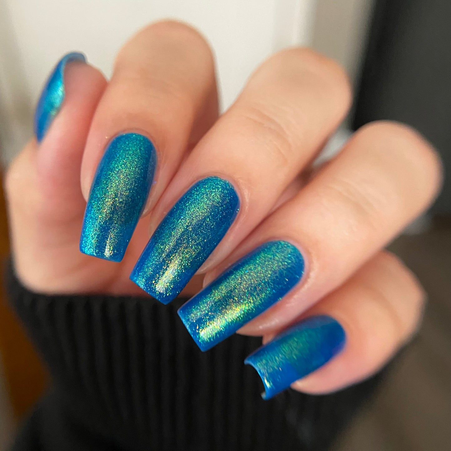 A close-up of a hand with manicured nails painted in a shimmering metallic blue nail polish with an iridescent green shift, wearing a black ribbed sweater.