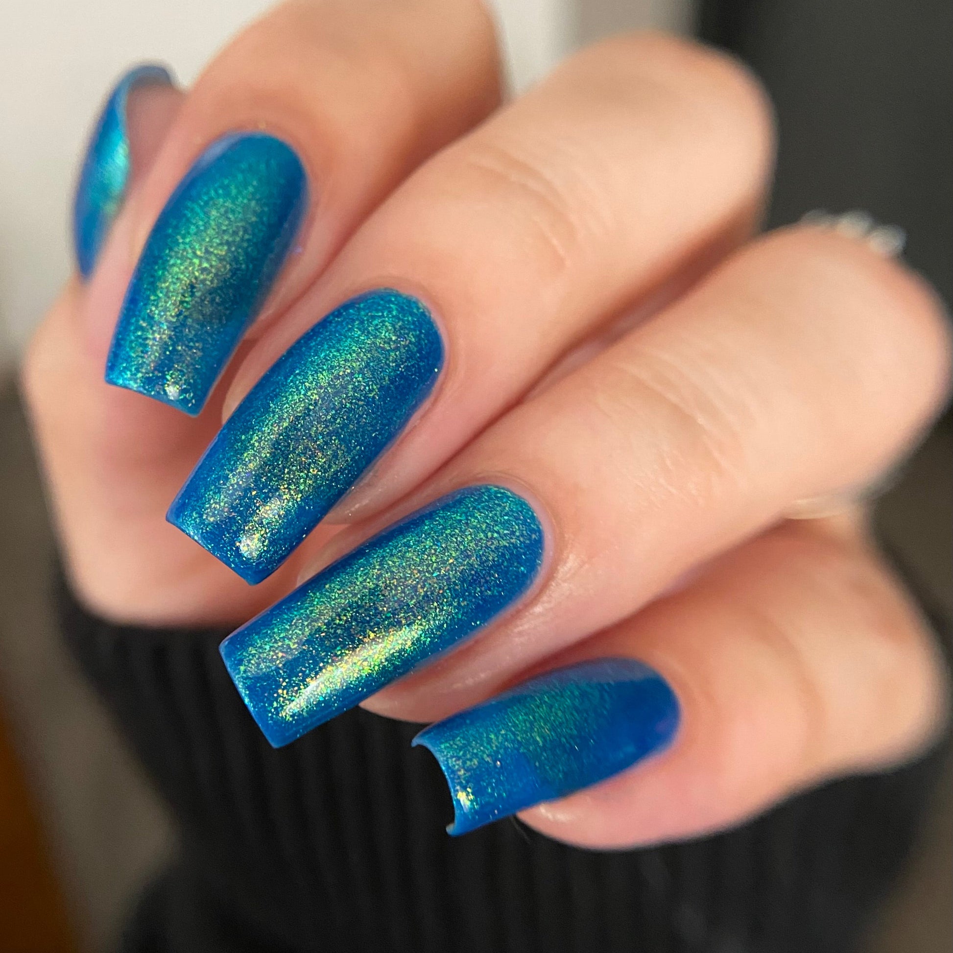 A close-up of a hand with long, almond-shaped nails painted in a shimmering blue nail polish that shifts with iridescent green and gold tones.