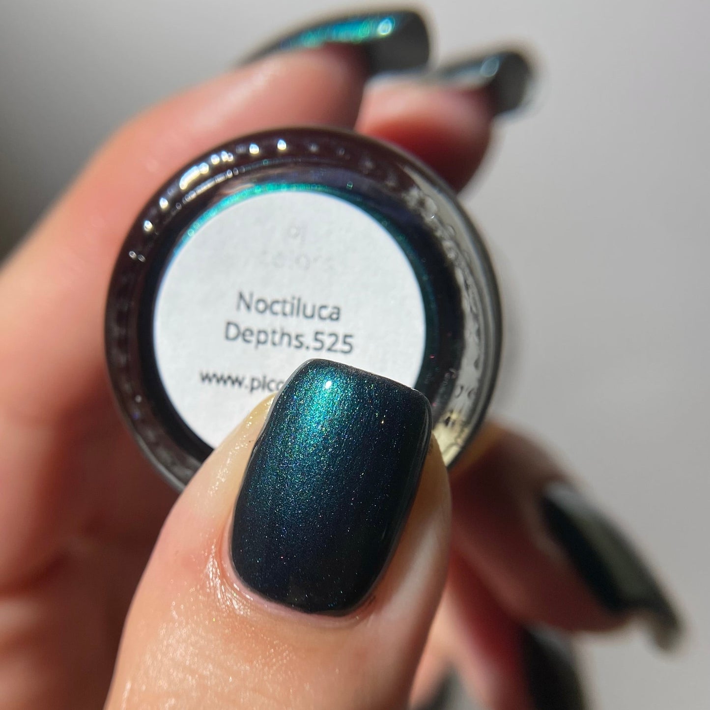 A close-up of a hand holding a small round jar of nail polish, showing the shade label 'Noctiluca Depths.525' and a glossy dark blue-green iridescent nail polish applied to the thumb.