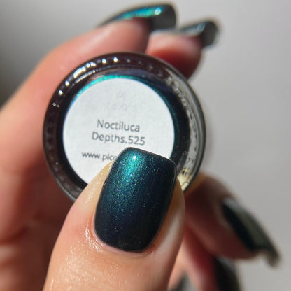 A close-up of a hand holding a small round jar of nail polish, showing the shade label 'Noctiluca Depths.525' and a glossy dark blue-green iridescent nail polish applied to the thumb.