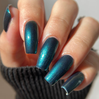 A close-up of a hand with long, almond-shaped nails painted in a metallic, shimmering teal-blue nail polish, wearing a dark ribbed knit sleeve.