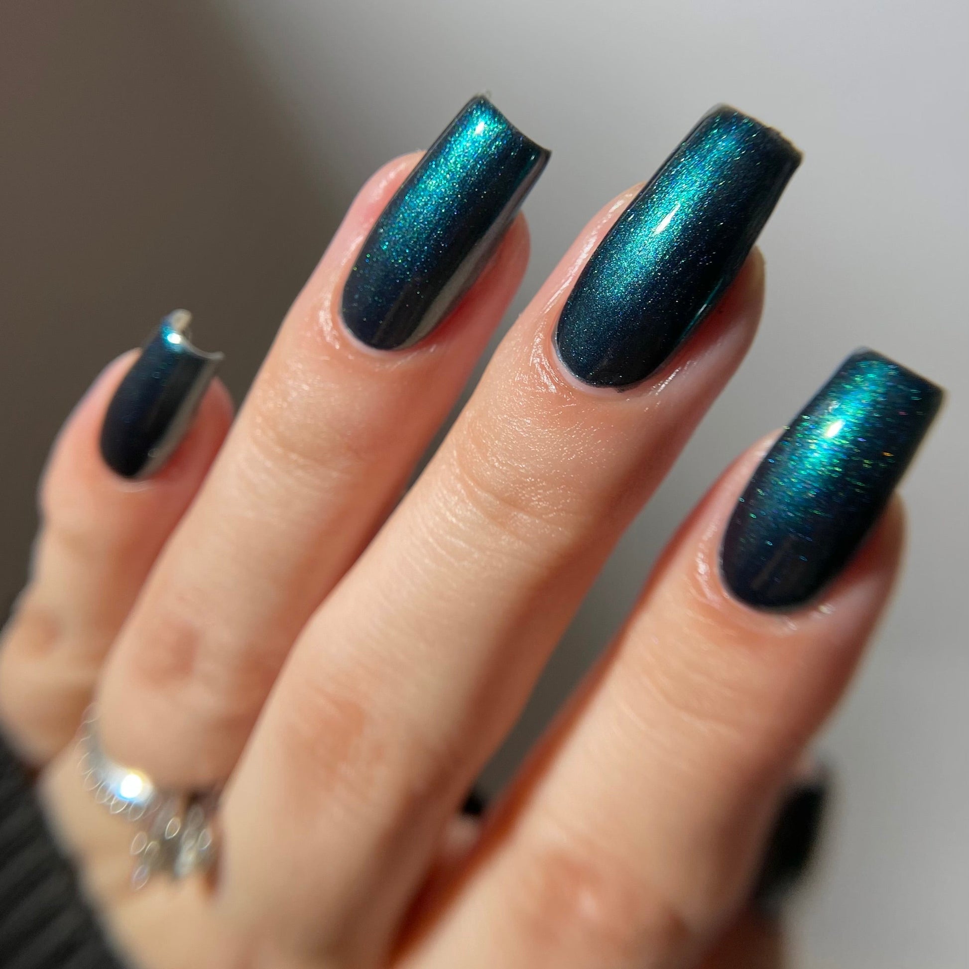 A close-up of a hand with long, coffin-shaped nails painted in a metallic teal-blue nail polish with shimmering iridescent flecks.