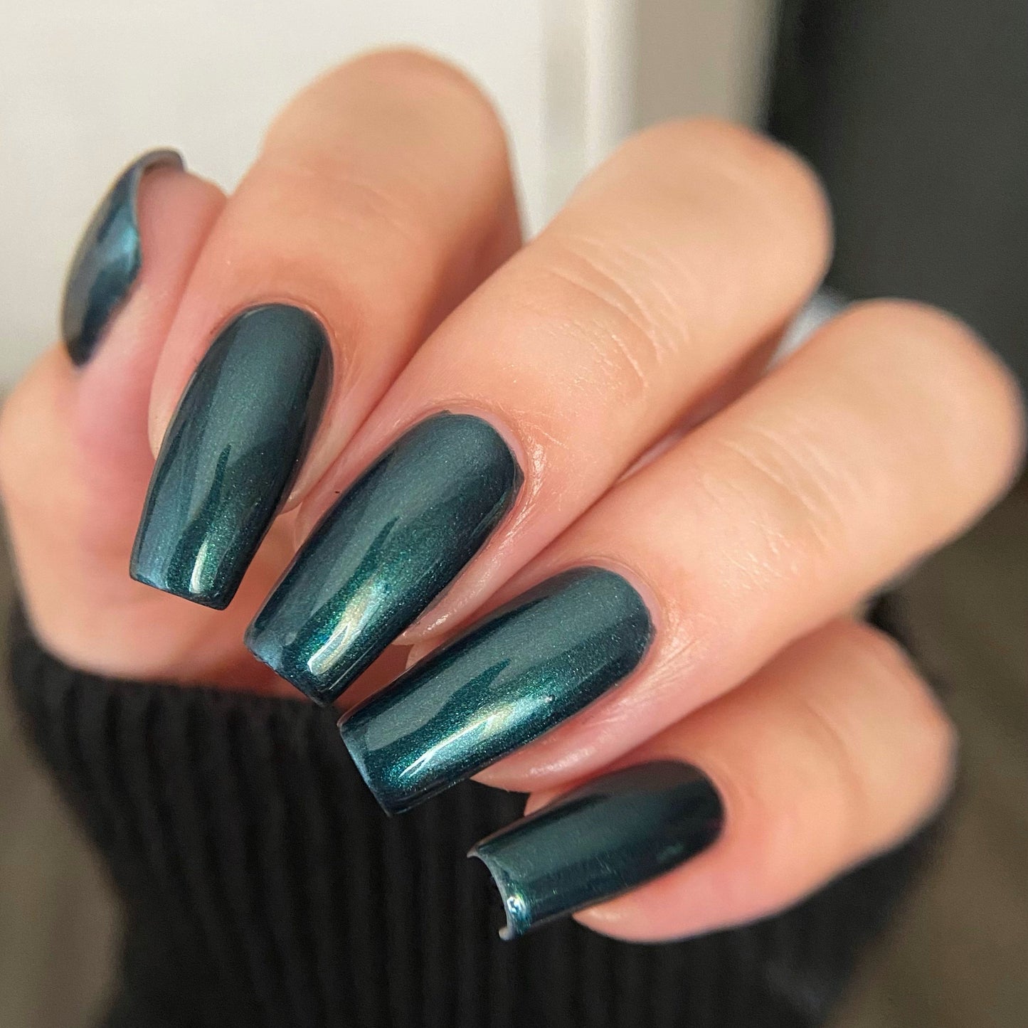 A close-up of a hand with long, almond-shaped nails painted in a metallic, shimmery dark teal green nail polish.
