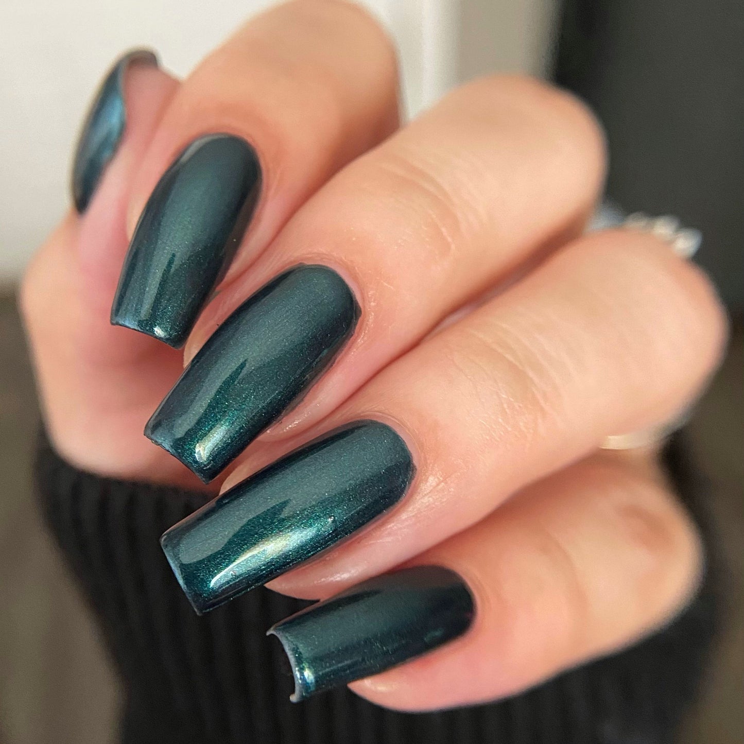A close-up of a hand with long, almond-shaped nails painted in a glossy dark teal green nail polish, wearing a black ribbed sweater.