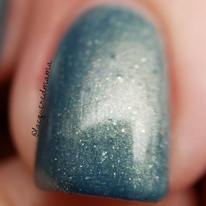 A close-up of a fingernail adorned with PI Colors' Sea Kelp.092 Green Nail Polish showcases its shimmering teal hue. This handmade nail lacquer features a subtle glitter effect and iridescent quality, with the background softly blurred.