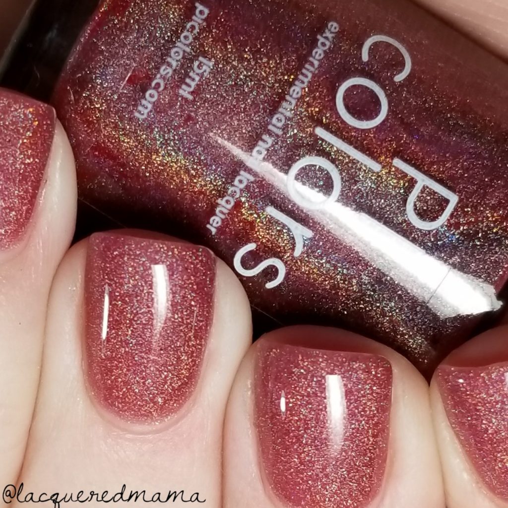 Fingers with nails painted in a sparkly, linear holo rose-gold polish hold a bottle of PI Colors nail color, "Rheum.011 Red Nail Polish," which has a similar shimmer. The 10-free polish bottle is labeled "PI Colors.