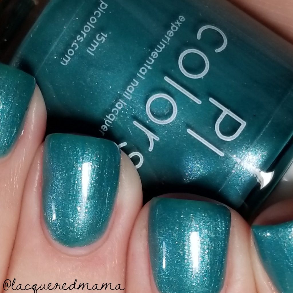 Close-up of fingernails painted with PI Colors' PavoC.023, a shimmery peacock green metallic nail polish. The 10-free PI Colors bottle, with its brand label prominently displayed in the background, highlights the same vibrant hue.