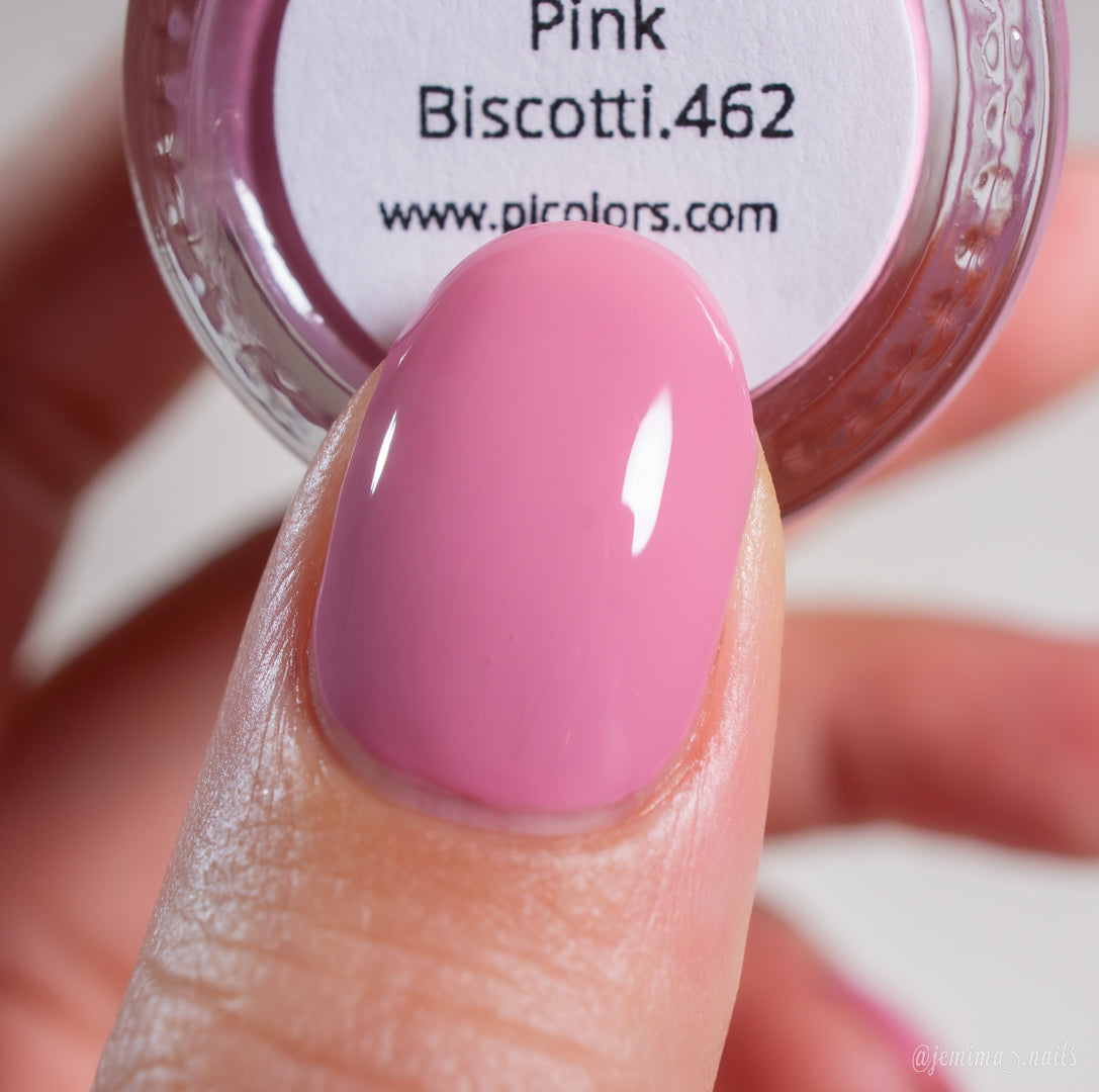 A close-up of a fingernail painted with glossy brownish pink nail polish. In the background is a partially visible bottle of PI Colors Pink Biscotti.462 nail polish, featuring a creme finish.