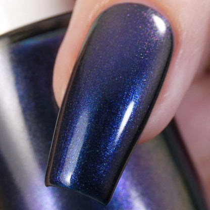 A close-up of a fingernail adorned with Judge None Choose One.000, an iridescent dark blue nail polish by PI Colors. It features a glossy multichrome finish with hints of purple and shimmer, set against an out-of-focus background that emphasizes the vibrant color.