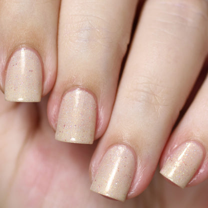 A close-up of square-shaped nails, expertly manicured with PI Colors' Shark's Eye Moonshell.062 Nude Cream Nail Polish, features a shimmery sheer beige hue complemented by subtle holographic glitter. The glossy finish highlights four fingers resting elegantly against each other.