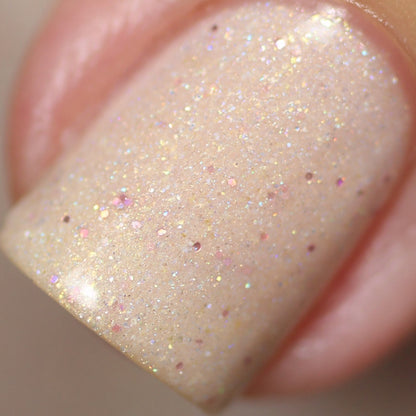 Close-up of a fingernail adorned with Shark's Eye Moonshell.062 Nude Cream Nail Polish from PI Colors. This handmade polish showcases multicolored sparkles, creating a subtle iridescent effect. The lighting accentuates the glossy finish and intricate texture of the holographic glitter.