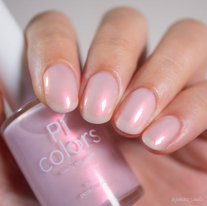 A hand with neatly manicured nails painted in PI Colors Snickerdoodle.405, a sheer, shimmery pale pink lacquer, holds the matching bottle. The nails have a glossy, iridescent finish.