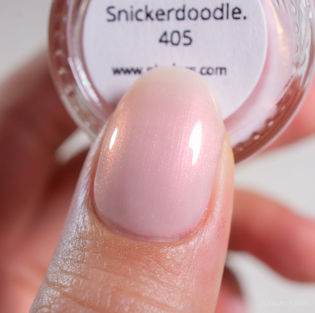 A close-up of a fingernail painted with PI Colors' Snickerdoodle.405, a handmade sheer, shimmery pale pink nail polish with a soft, glossy finish for everyday luxury. The bottle label reads "Snickerdoodle.405.