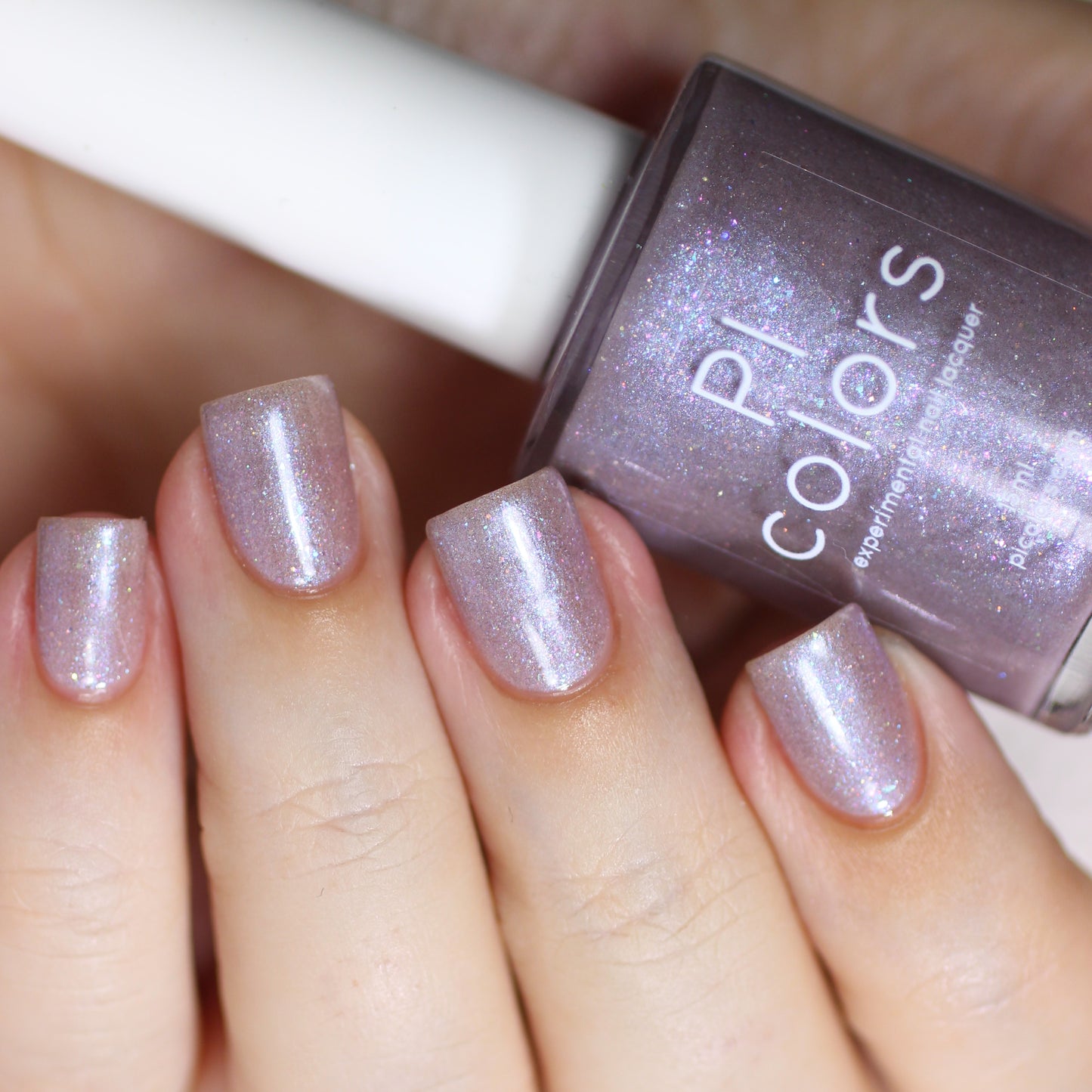 A close-up shot showcases a hand with nails adorned in a shimmery lavender polish, featuring holographic glitter. The fingers are gripping a bottle of PI Colors' Snow Cherries From France.101 Purple Nail Polish, which provides a glossy finish and dazzling effect to elevate your manicure to new heights.