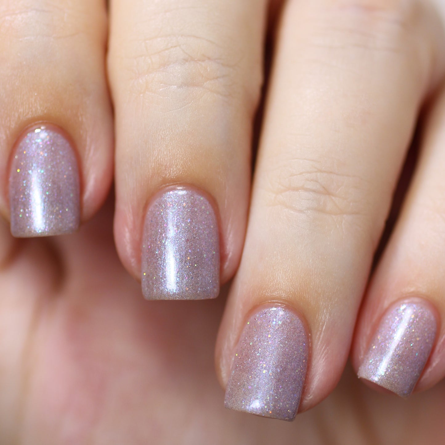 A hand with short, square-shaped nails is painted in the shimmering light lavender shade of Snow Cherries From France by PI Colors, featuring holographic glitter. The neatly manicured nails sparkle elegantly against the fair skin tone.