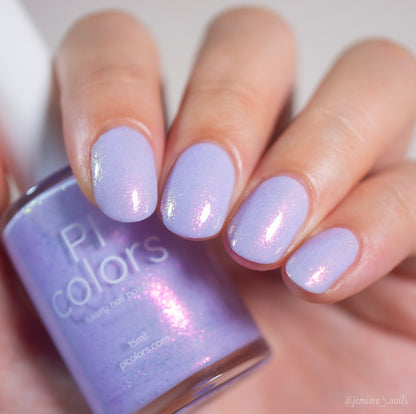 A hand with neatly manicured nails painted in Snowball.485, a light purple handmade polish by PI Colors, holds a matching bottle. The polish has a subtle iridescent pink sheen for an elegant finish.