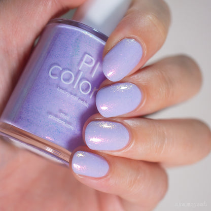 A hand holds a bottle of PI Colors Snowball.485 handmade shimmer nail polish in pastel purple, with freshly painted nails displaying the same shimmery lavender hue.