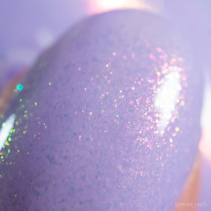 A close-up of a fingernail painted with PI Colors’ Snowball.485, a shimmery light purple nail polish with iridescent glitter that reflects pink, green, and purple sparkles under the light.