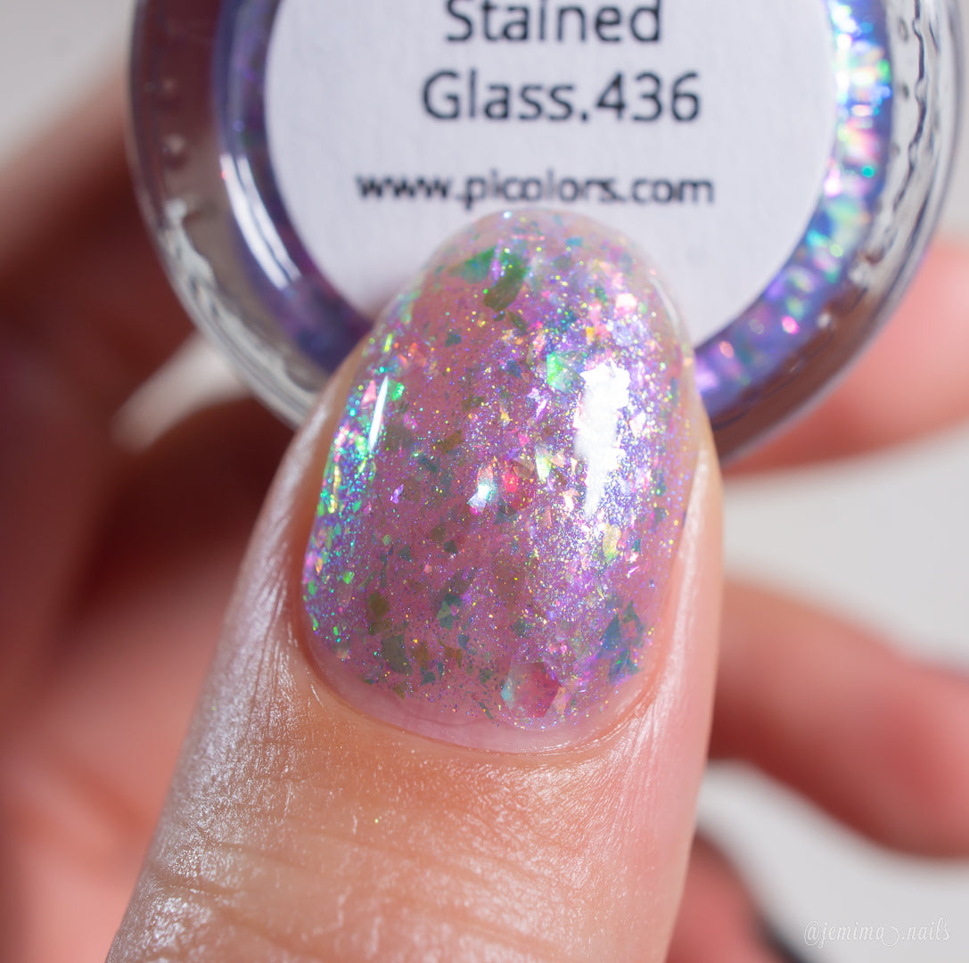 A close-up of a fingernail with shiny, iridescent flakie polish featuring pink, purple, and green flakes. In the background, a jar of PI Colors handmade nail polish labeled "Stained Glass.436" is partially visible.