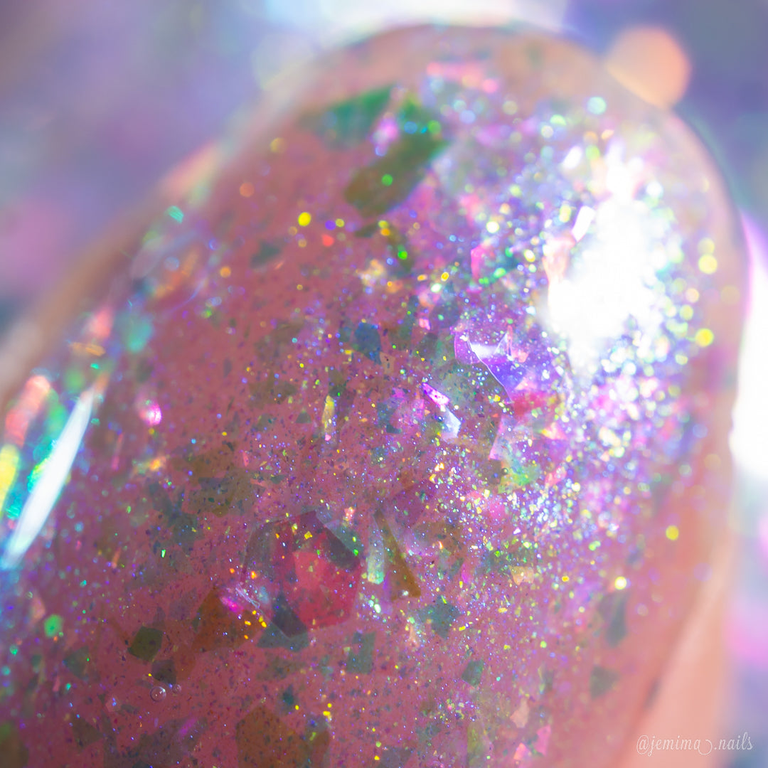 A close-up of a fingernail displays PI Colors' Stained Glass.436, a handmade flakie nail polish with glossy finish and multicolored glitter flakes reflecting pink, purple, green, and gold hues under bright light.