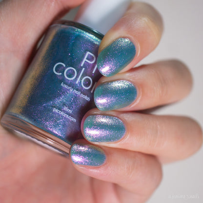 A hand with short, rounded nails painted in PI Colors Sugar Sparkles.451, a shiny iridescent blue-green nail lacquer with pinkish-purple shimmer, holds a matching bottle of polish.