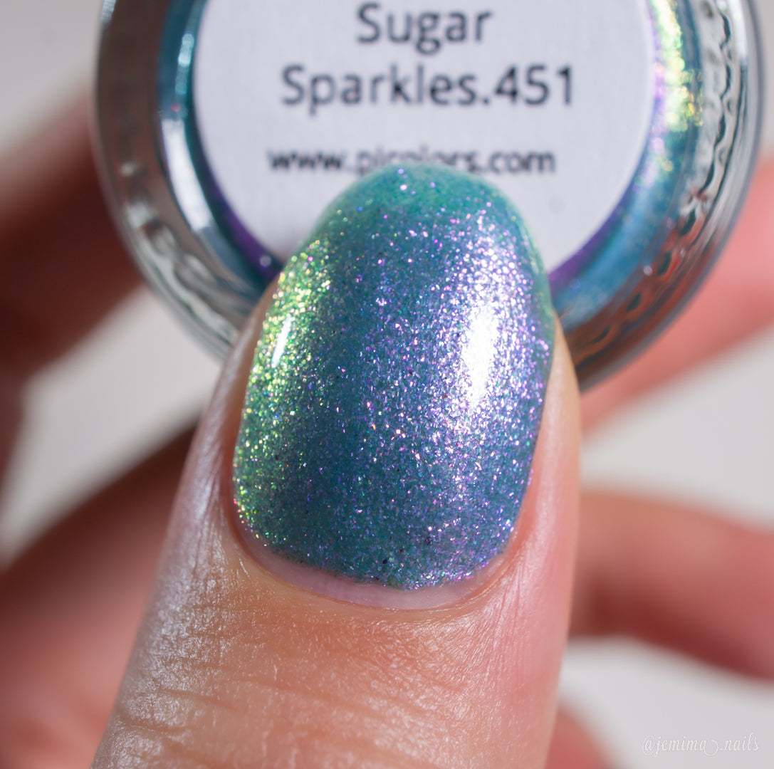 A close-up of a thumb adorned with glittery, iridescent blue and purple luxury nail lacquer. In the background, a PI Colors Sugar Sparkles.451 nail polish bottle is partially visible.