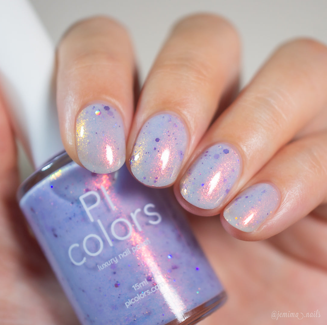 A hand with neatly manicured nails painted in Thistle Plum.100, a shimmery pale purple polish with pink, gold, and holographic violet glitter, holds the matching PI Colors bottle labeled "Thistle Plum.100" next to the nails.