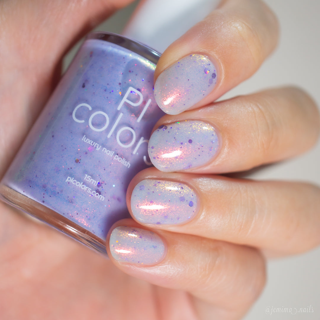 A hand with short, rounded nails painted in sheer lilac "Thistle Plum.100" by PI Colors—with pink and holographic violet glitter—holds a matching PI Colors luxury nail polish bottle.