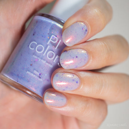 A hand with short, rounded nails painted in sheer lilac "Thistle Plum.100" by PI Colors—with pink and holographic violet glitter—holds a matching PI Colors luxury nail polish bottle.