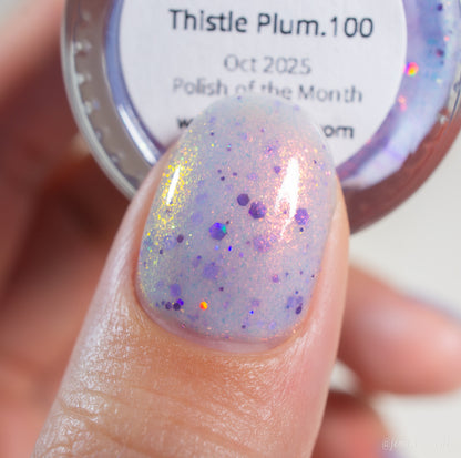 A close-up of a fingernail painted with PI Colors’ Thistle Plum.100, a sheer pale purple polish with gold iridescent shimmer and holographic violet glitter. A handmade bottle labeled "Thistle Plum.100" is blurred in the background.