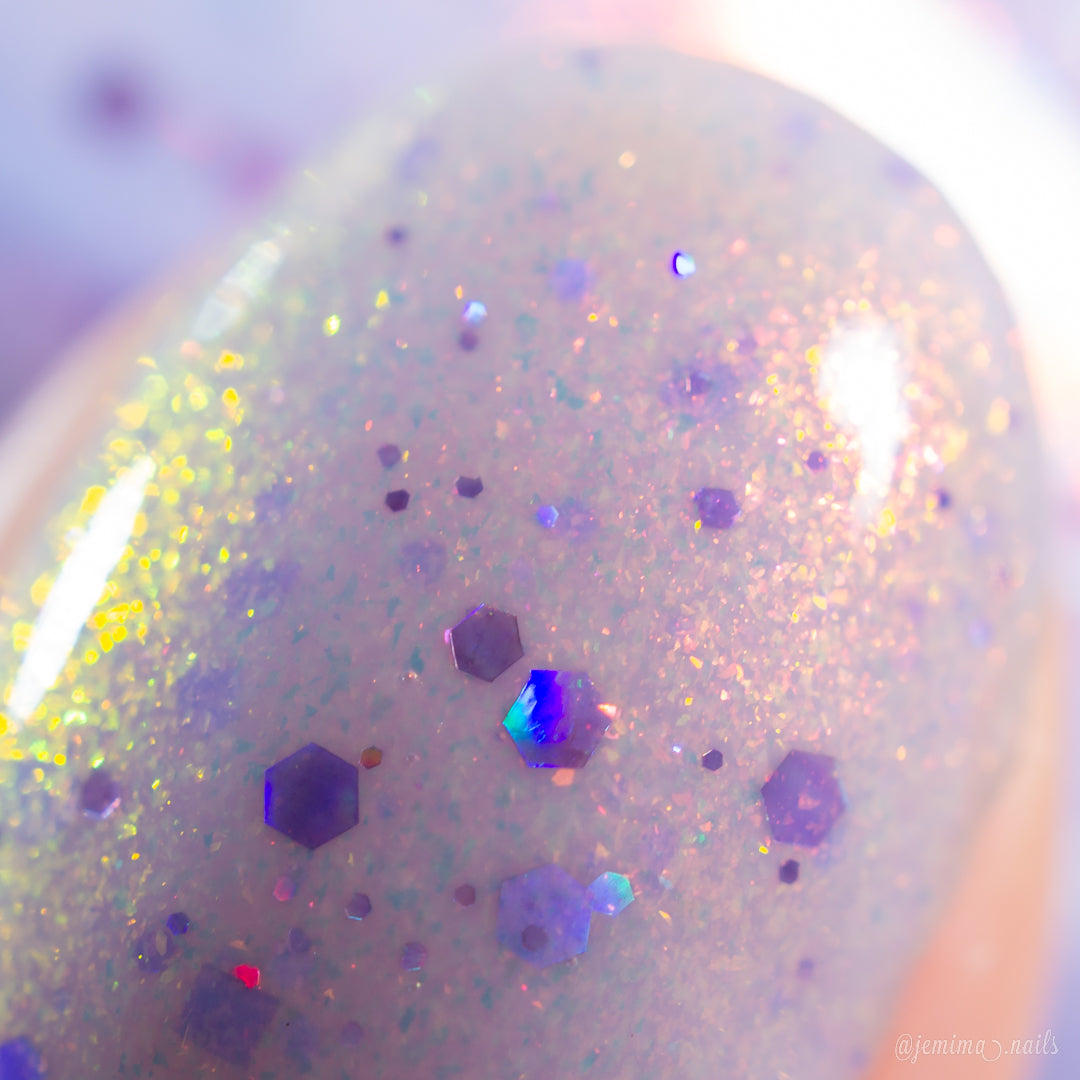A close-up of a fingernail wearing PI Colors Thistle Plum.100, a handmade nail polish in glossy, iridescent pale purple with hexagonal and holographic glitter in blue, purple, and gold for a pastel, light-reflecting look.