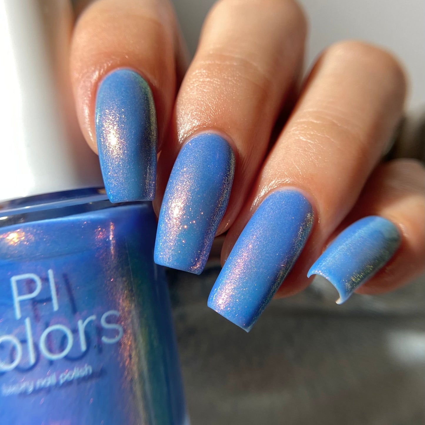 A close-up of a hand with long, glossy blue nails wearing a shimmering blue nail polish, with the matching blue bottle partially visible in the foreground. The brand text 'PI colors' is visible on the bottle.