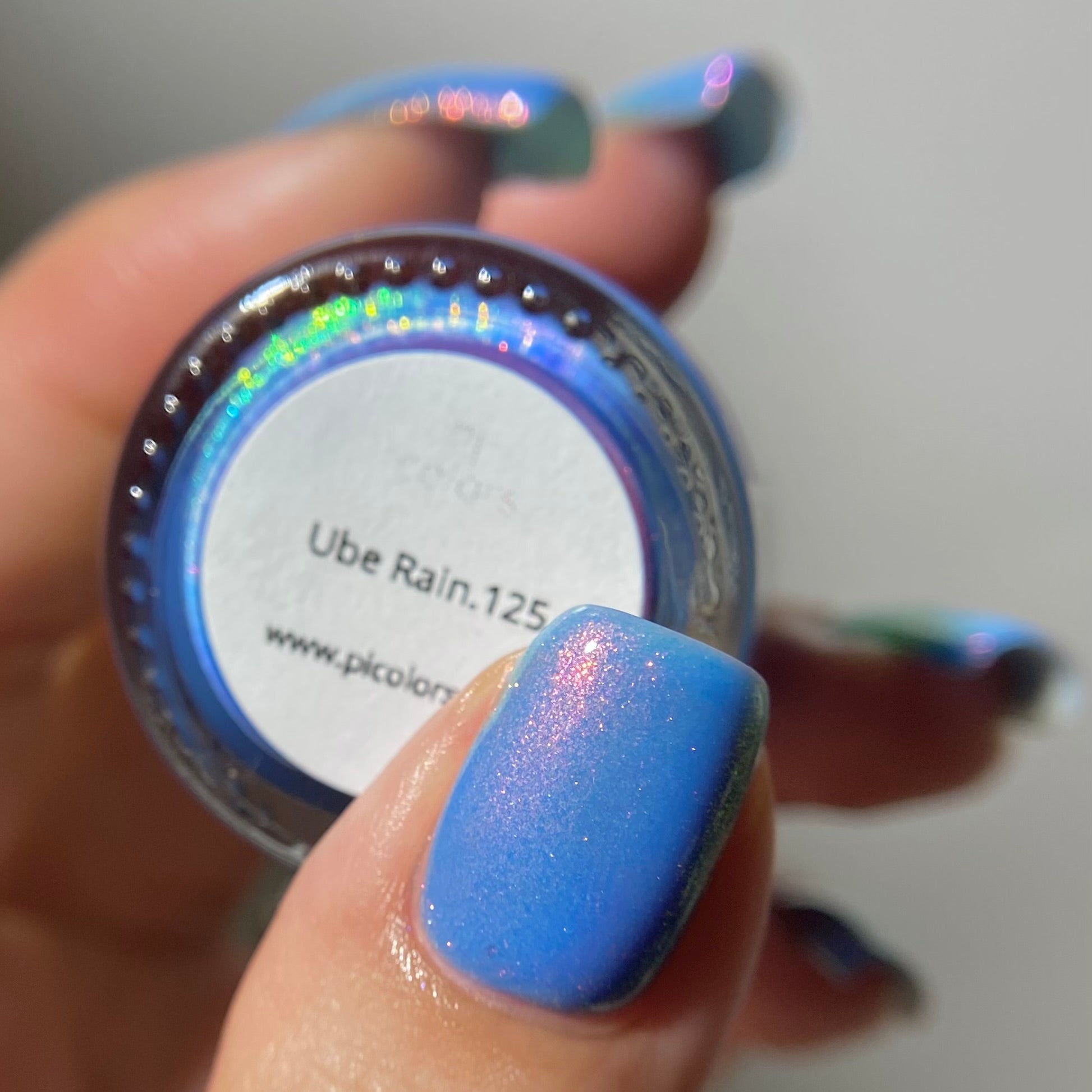 A close-up of a hand holding a small round bottle of nail polish. The nail polish on the fingernail and in the bottle shows a holographic blue shimmer that shifts with light, labeled 'Ube Rain.