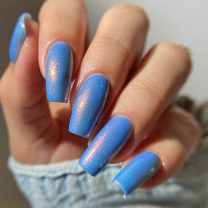 A close-up of a hand with almond-shaped nails painted in a glossy medium blue with a subtle iridescent shimmer, wearing a light denim sleeve.