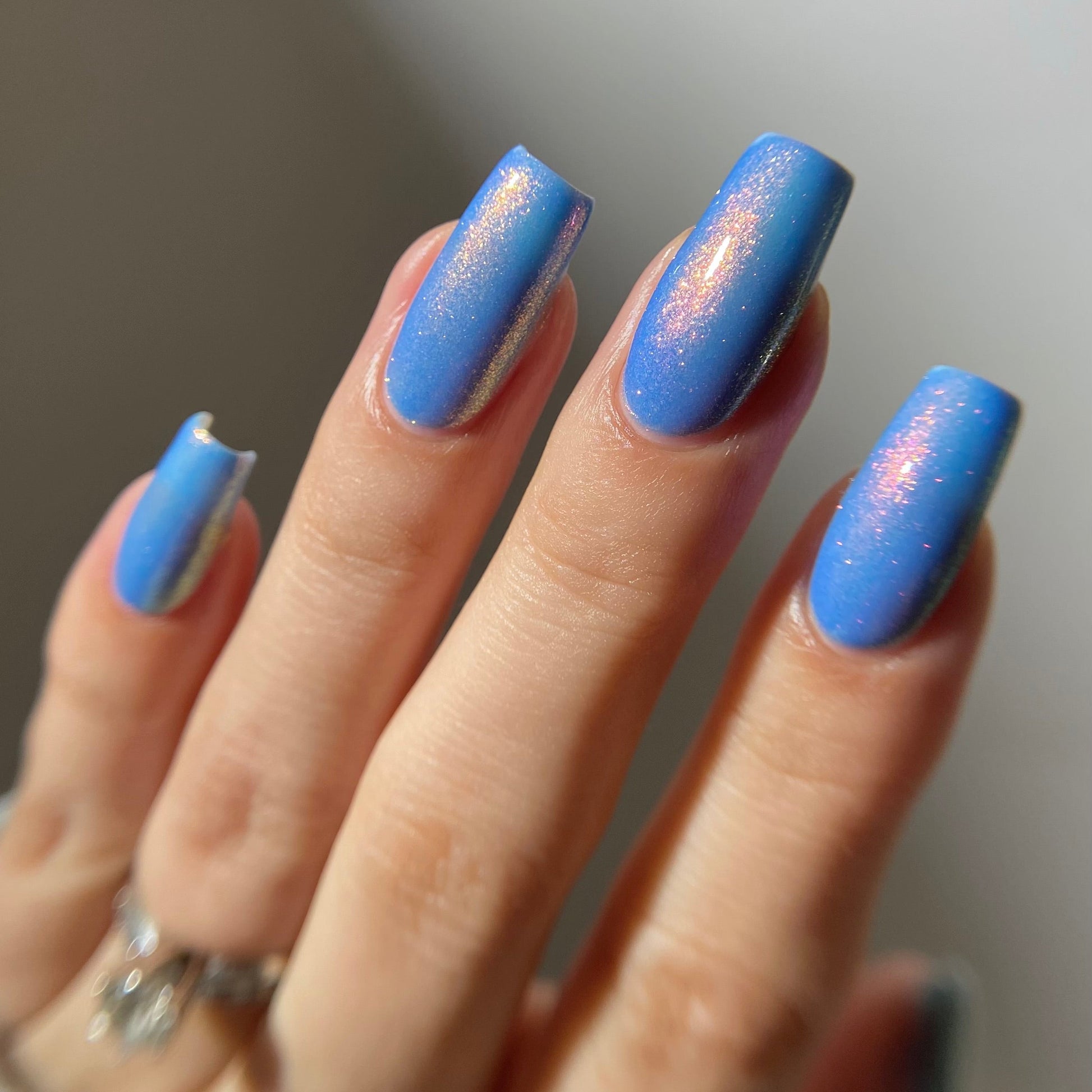 A close-up of a hand with medium blue nails featuring a shimmering, iridescent finish. The hand is shown against a soft, out-of-focus neutral background.