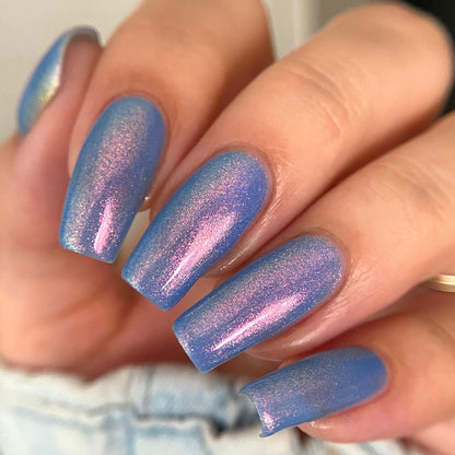 A close-up of a hand with long, almond-shaped nails painted in a shimmering iridescent blue polish that shifts to lavender and pink. The hand is resting near light-wash denim jeans, with a gold ring visible on one finger.