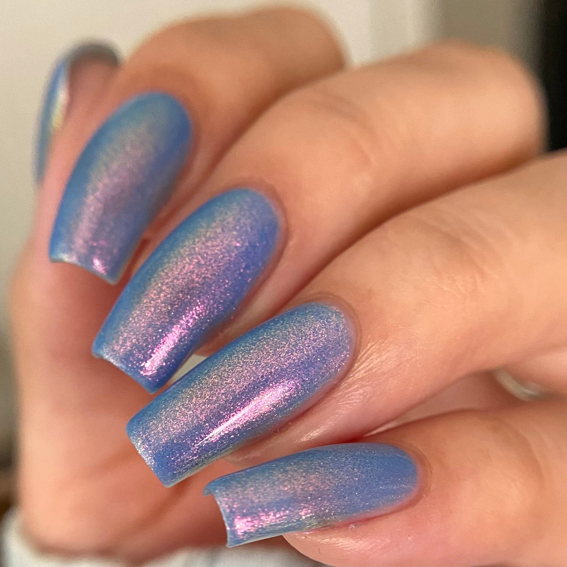 A close-up of a hand with long almond-shaped nails painted in a shimmering iridescent blue-to-purple holographic nail polish.