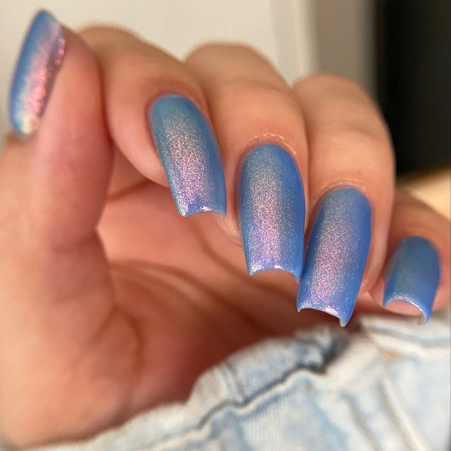 A close-up of a hand with manicured nails painted in a shimmering iridescent blue polish that shifts toward lavender and pink, resting against light denim fabric.