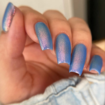 A close-up of a hand with manicured nails painted in a shimmering iridescent blue polish that shifts toward lavender and pink, resting against light denim fabric.