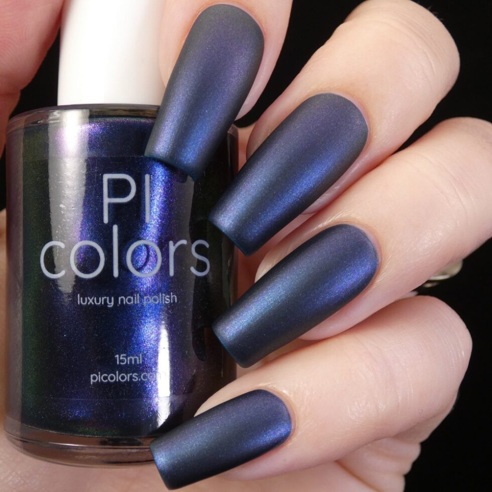 A close-up captures a hand with long, matte dark blue nails holding a bottle of PI Colors' Judge None Choose One.000 Luxury Nail Lacquer. The polish showcases a multichrome finish with shimmering, iridescent hints of blue and purple, evoking 90s nostalgia.