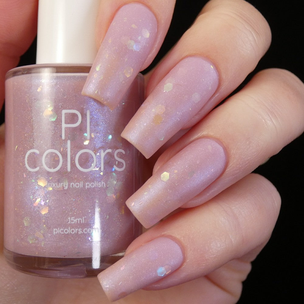 A hand with long, square-shaped nails painted in shimmery pastel pink holds a bottle of PI Colors' Special Week.000 Pink Nail Polish. The label reads "Special Week.000 Pink" and the bottle sparkles with iridescent glitter, capturing the essence of elegance and sophistication in each handmade drop.