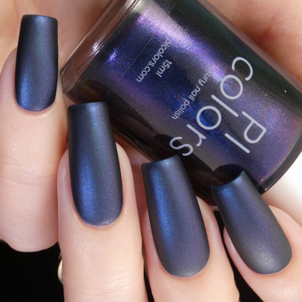 A hand with long nails painted in a dark blue, matte metallic polish exudes 90s nostalgia. The "Judge None Choose One.000" bottle from PI Colors is nestled between the fingers, revealing a shimmering iridescent effect and subtle multichrome finish against a dark background.