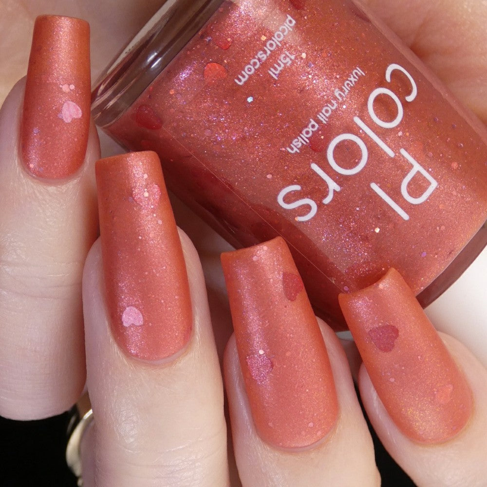 Close-up of hands with long, rectangular-shaped nails painted in a luxurious coral lacquer featuring heart-shaped glitter accents. Elegantly held between the fingers is a matching bottle of PI Colors Strawberry Jam.228 Red Nail Polish, complete with red glitter details.