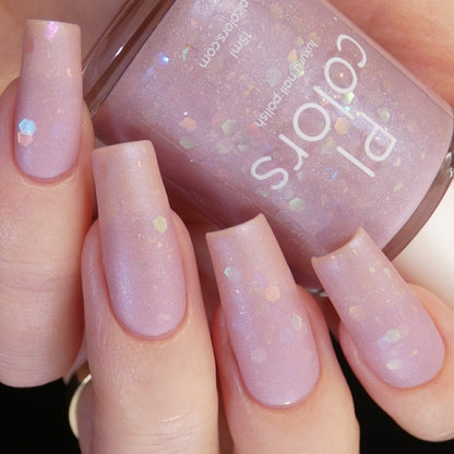 A hand showcasing long, square-shaped nails painted in a light pink hue with iridescent glitter holds a bottle of "Special Week.000 Pink Nail Polish" by PI Colors, capturing elegance and finesse.